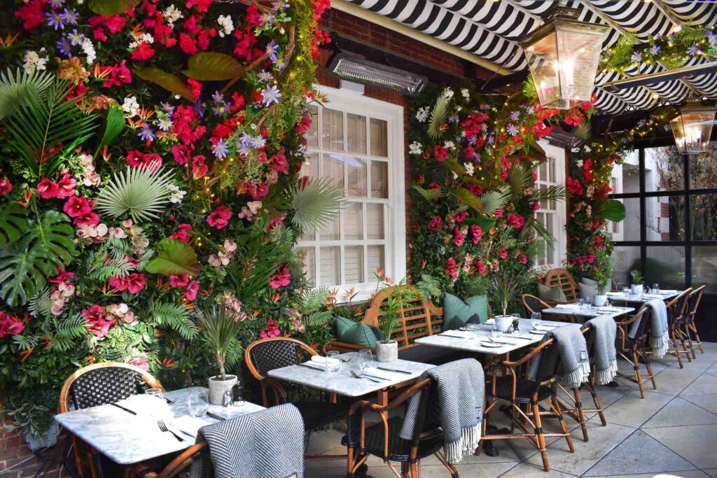 Adorable restaurante al aire libre con paredes de flores brillantes y mantas sobre las sillas de mimbre para mantener a los clientes calientes junto con un calentador Bromic